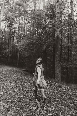 Black and white portrait of young woman in dress laughing on leaf-covered forest path in fall.