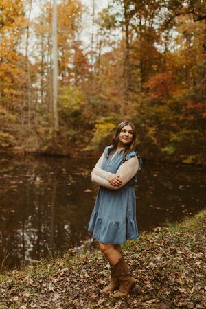 Young woman in blue denim dress and boots standing by autumn forest pond with colorful fall foliage.