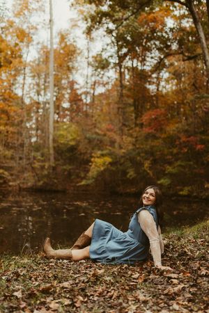 Young woman in blue dress sitting by peaceful pond surrounded by colorful autumn trees and fallen leaves.