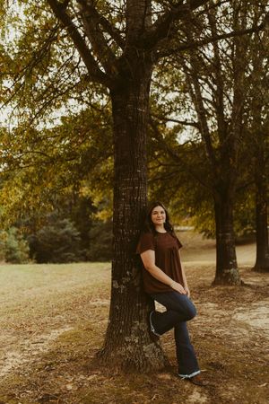 Young woman in brown top leaning against large tree trunk in golden autumn park setting.