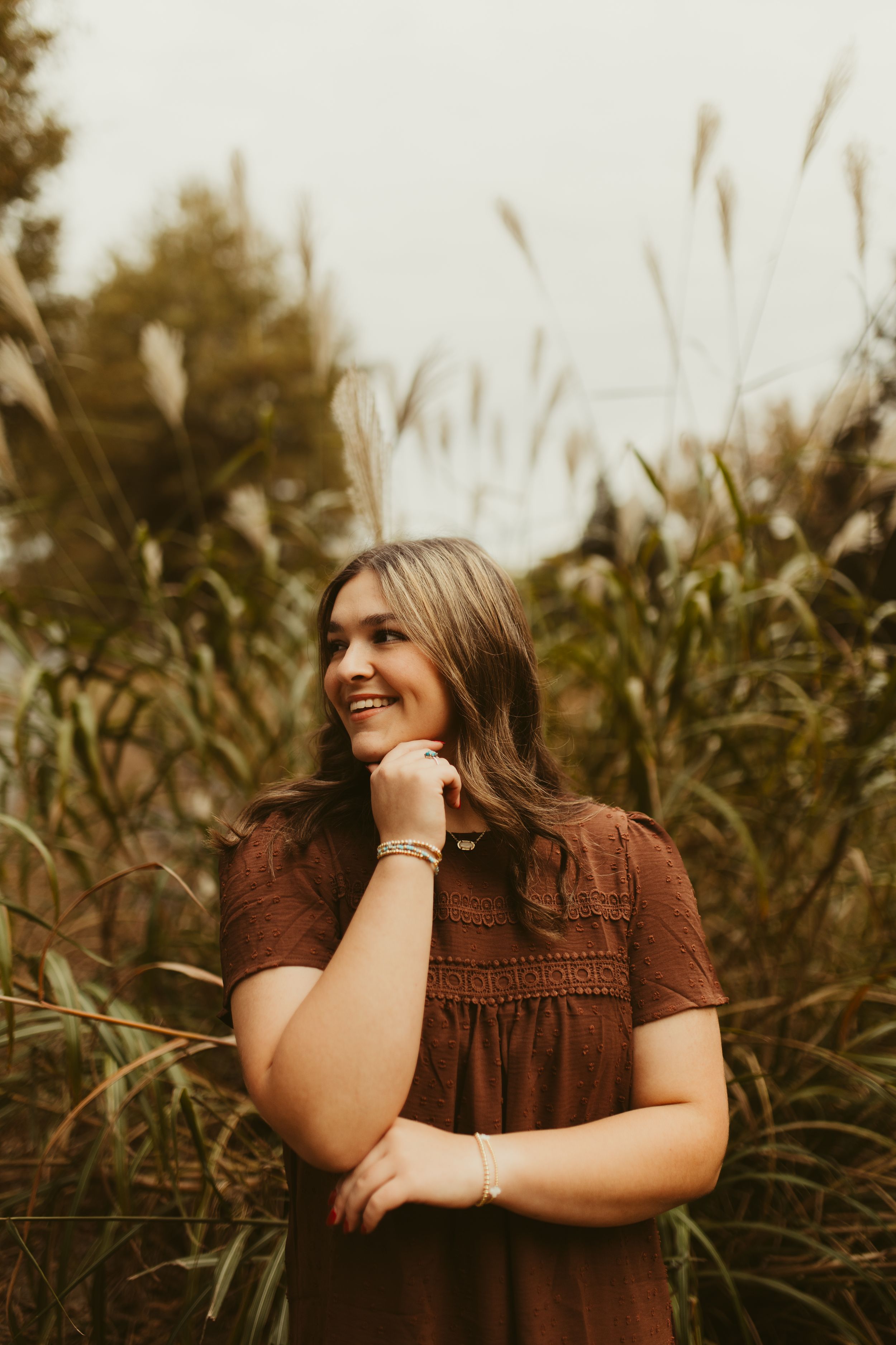 Smiling young woman in brown dress standing before tall ornamental grasses in warm outdoor portrait.
