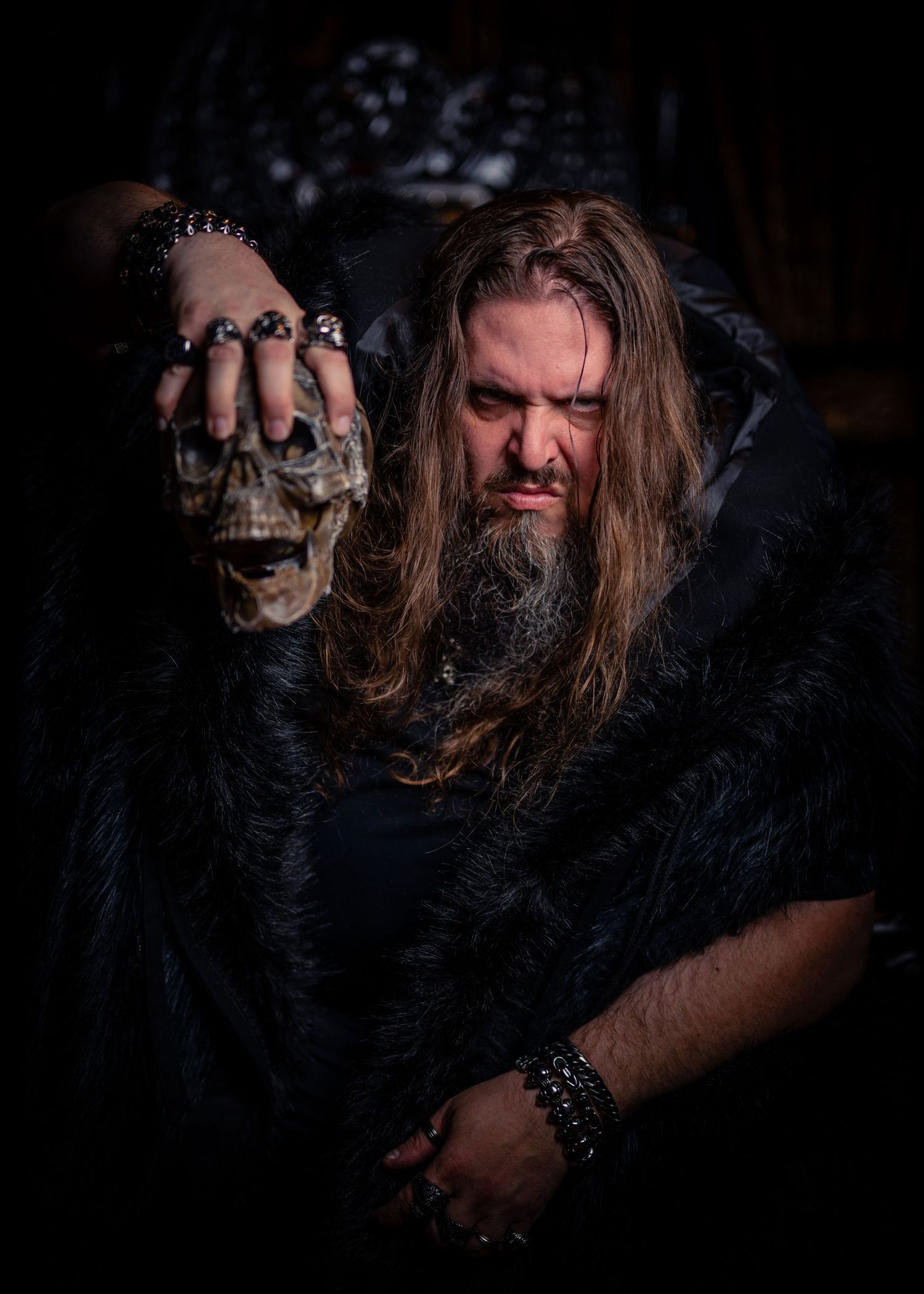 Dark atmospheric portrait featuring long-haired person in black clothing holding a decorative skull against shadowy backdrop.