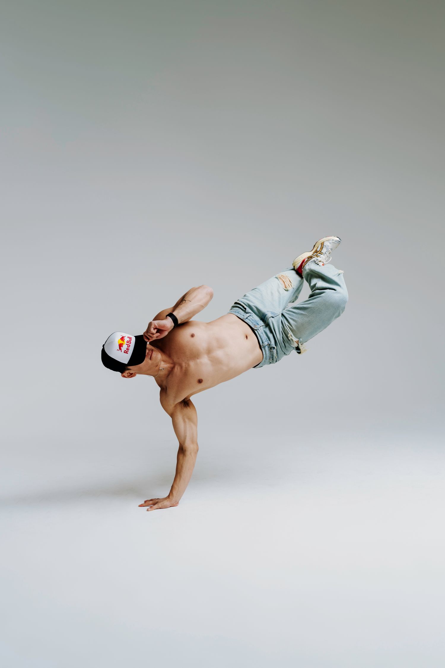 Shirtless dancer performing a breakdance freeze move on one arm while wearing jeans and a baseball cap in a studio.