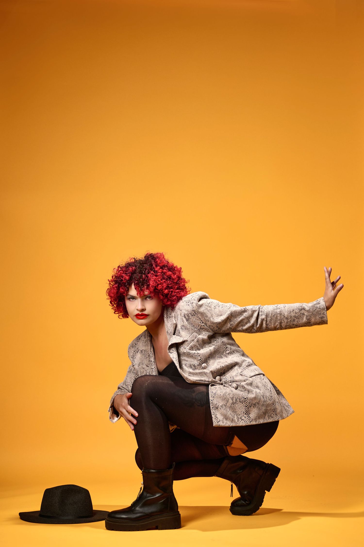 A performer in red curly wig and beige jacket crouches in a dynamic pose beside a black hat against an orange backdrop.