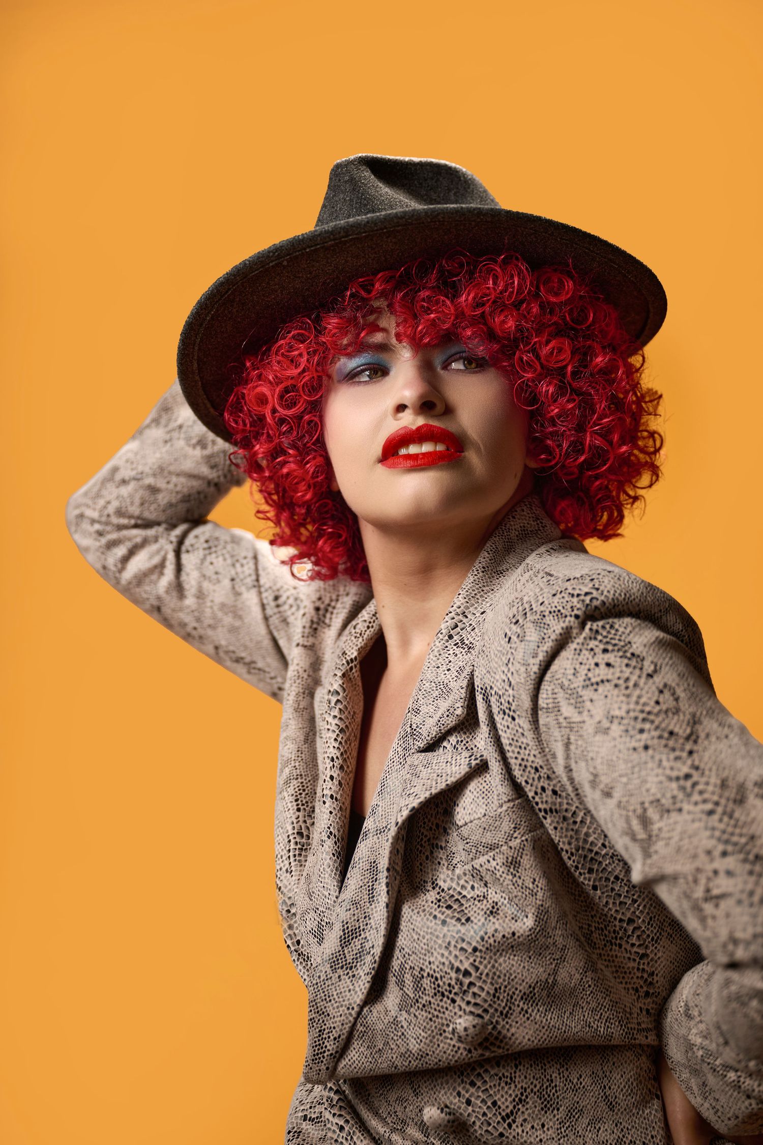 Fashion portrait with vibrant red curly hair wearing a snakeskin print blazer and black hat against an orange backdrop.