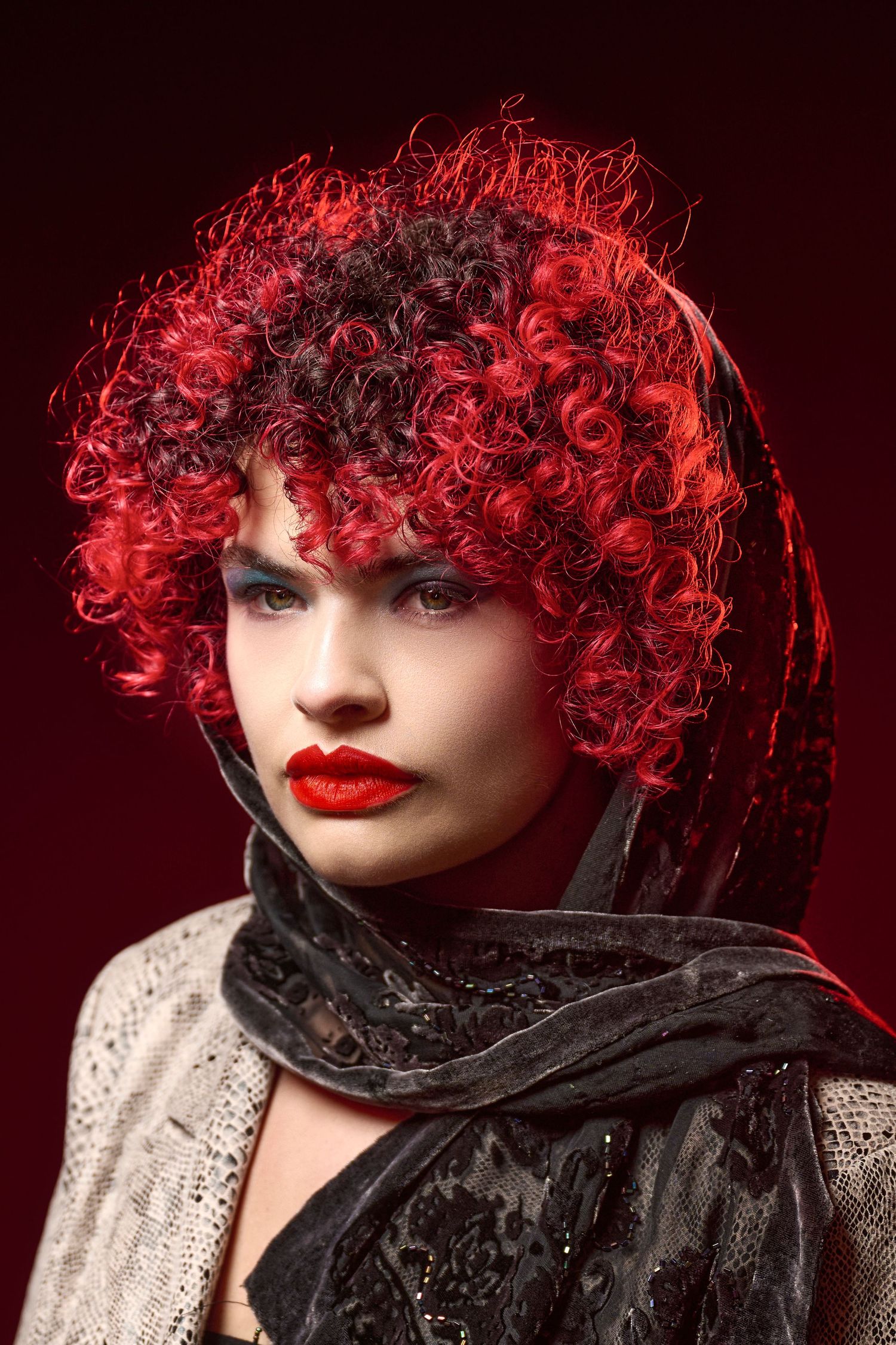 Fashion portrait with vibrant red curly hair, bold red lips, and dramatic black scarf against dark background.