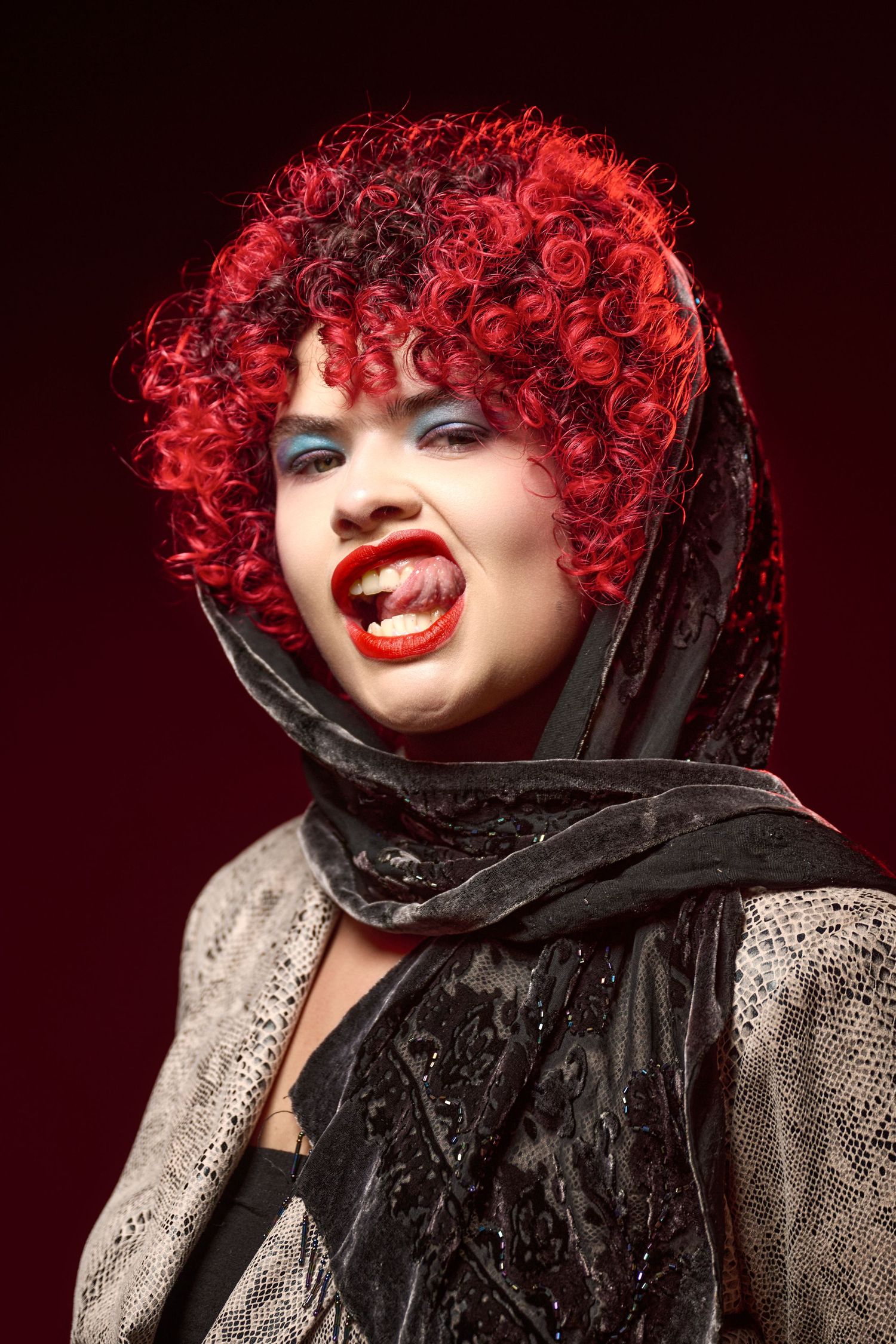 Dramatic portrait featuring bright red curly hair, red lipstick, black scarf and silver outfit against dark background.