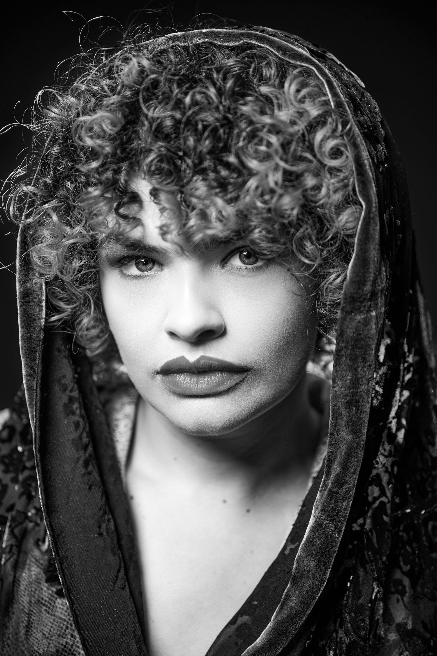 Dramatic black and white portrait of a figure wearing a dark veil with natural curly hair and defined features.