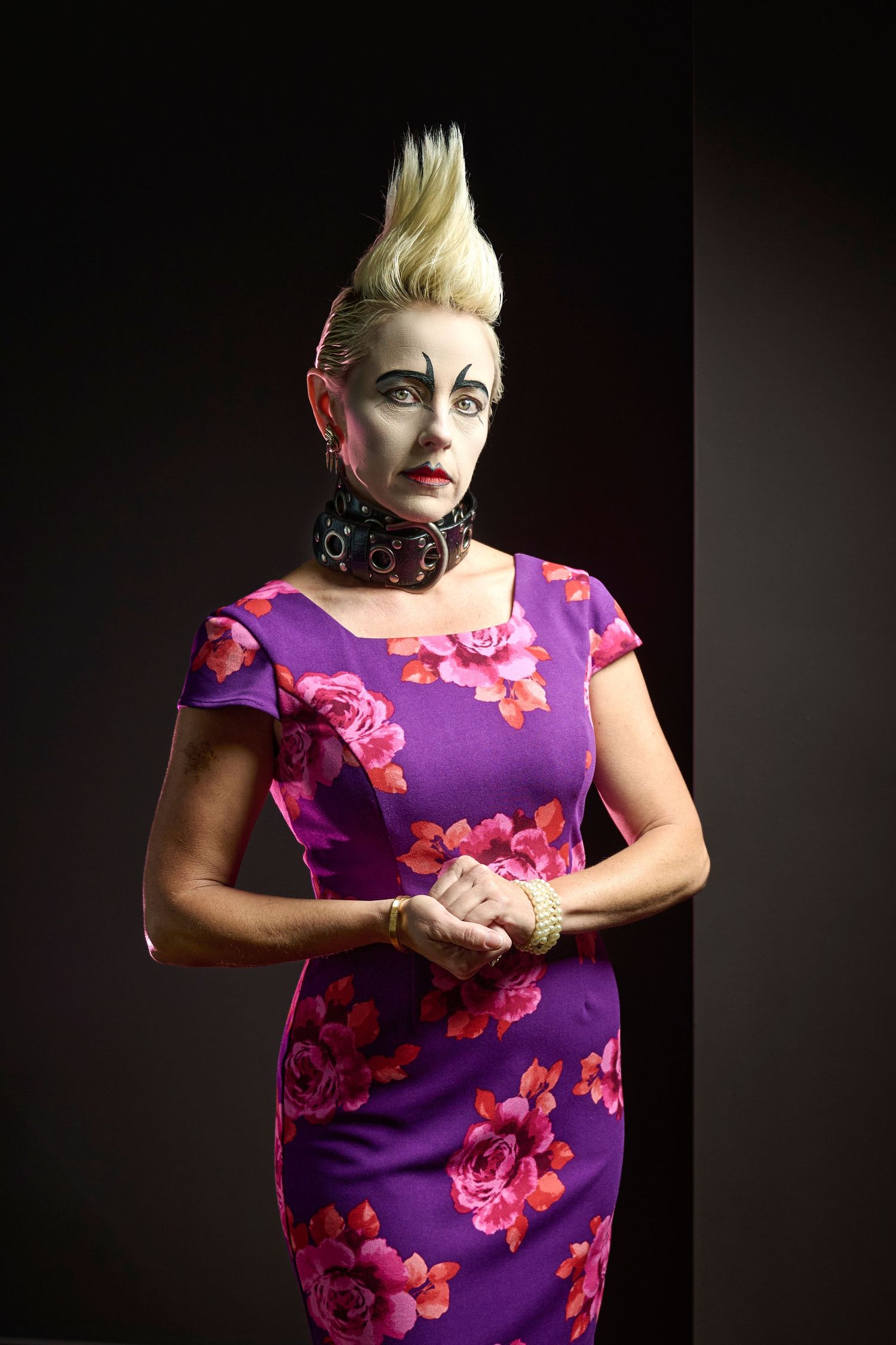 Dramatic portrait of figure in purple floral dress with spiked blonde hair and studded choker against dark background.