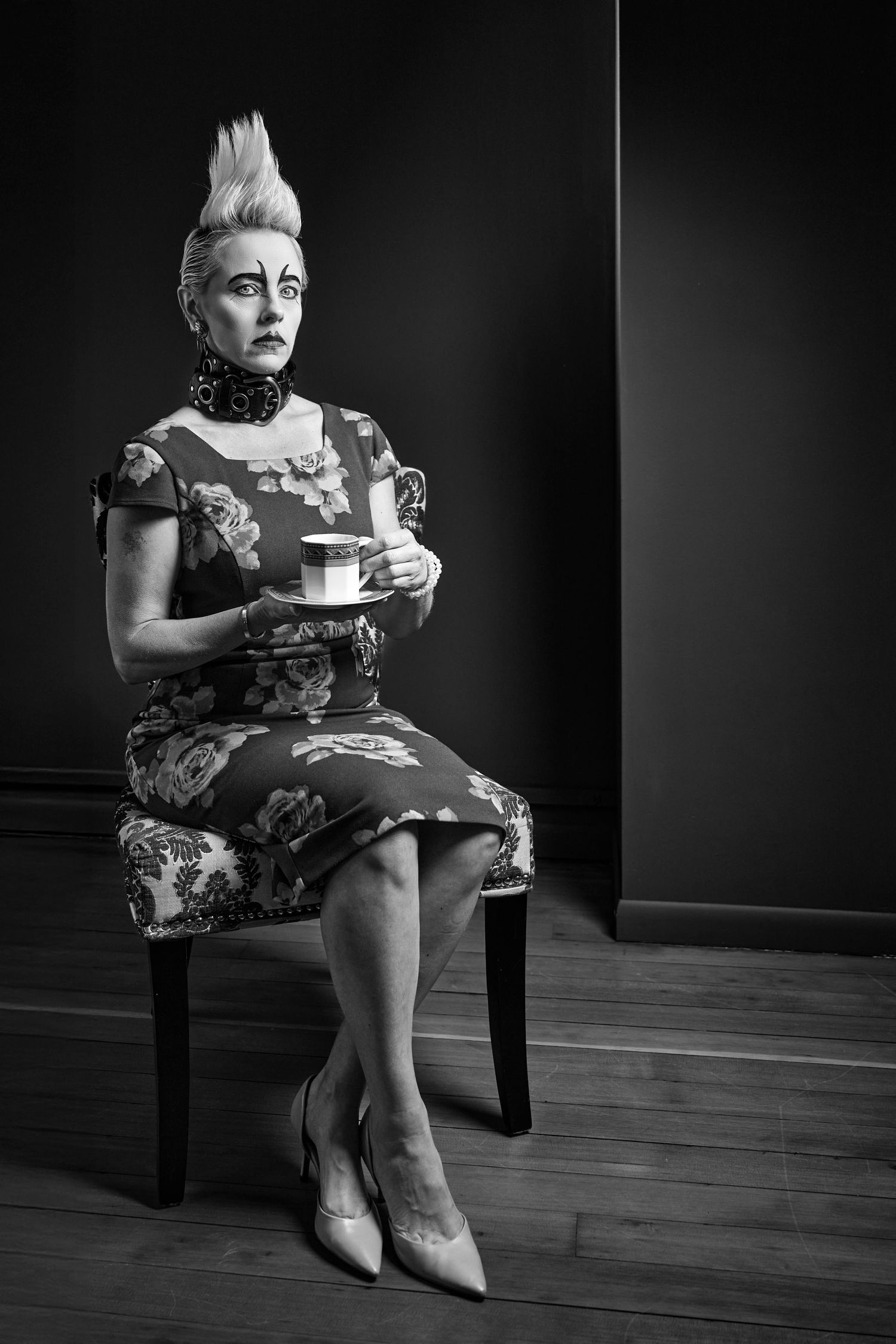 A dramatic black and white portrait of a person in vintage dress with mohawk hairstyle sitting formally with tea.