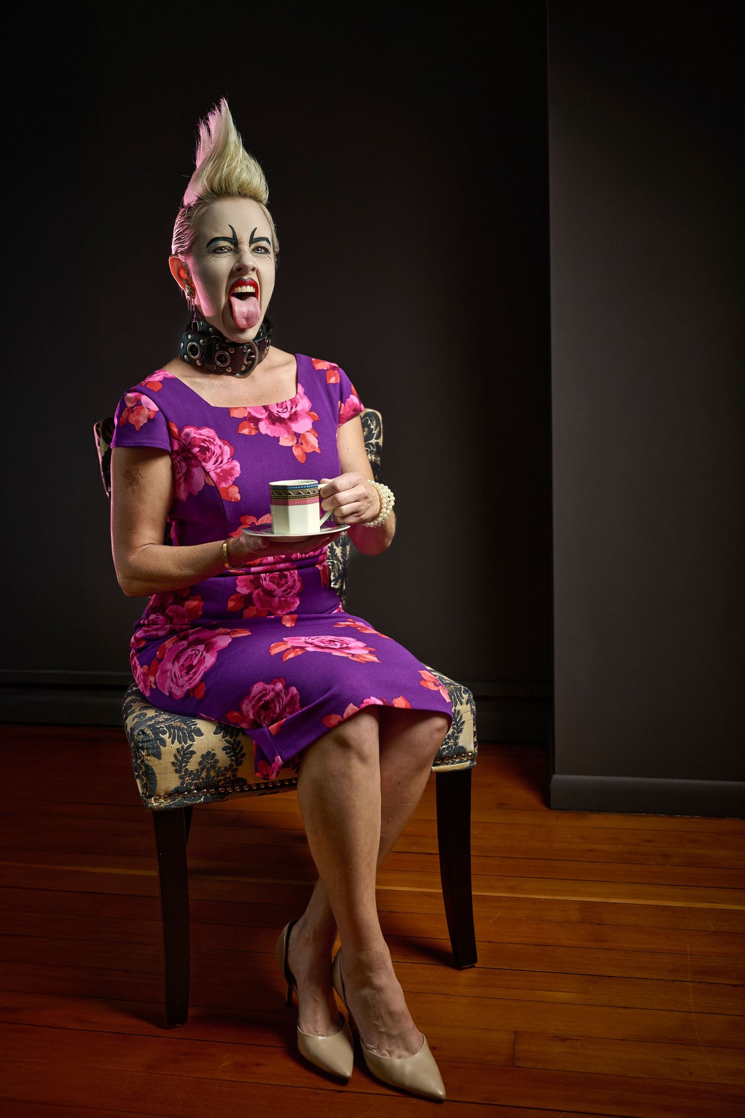 A person in a vibrant purple floral dress and blonde mohawk hairstyle sits on a chair holding a coffee cup.