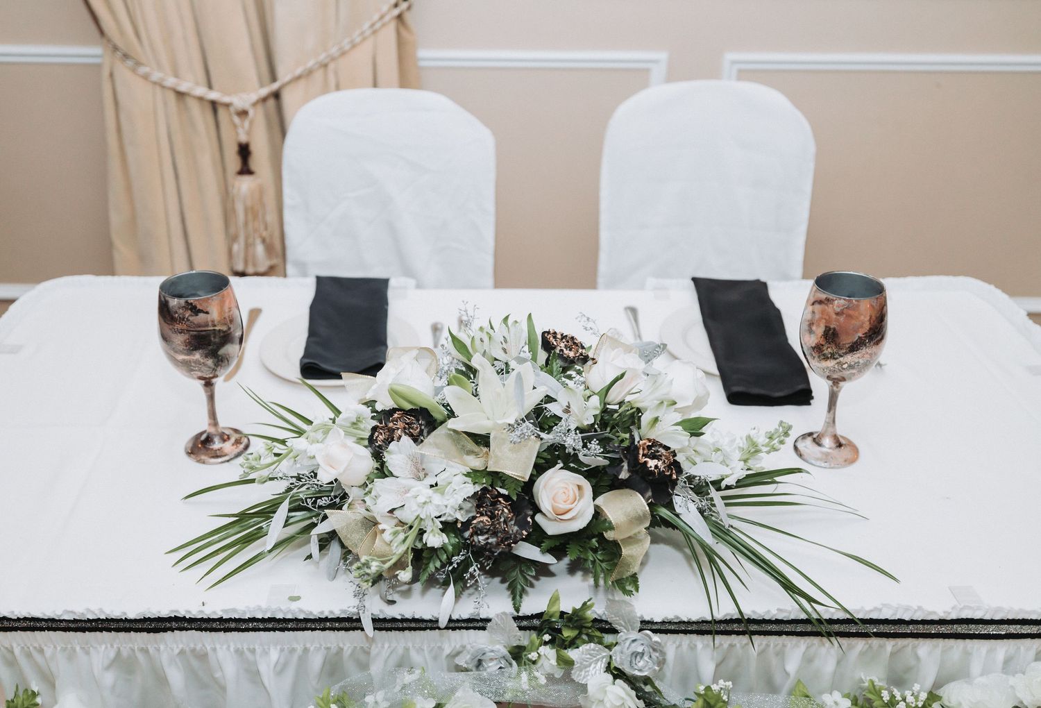 Elegant wedding table setting with white floral centerpiece, wine glasses, and black napkins on white tablecloth.