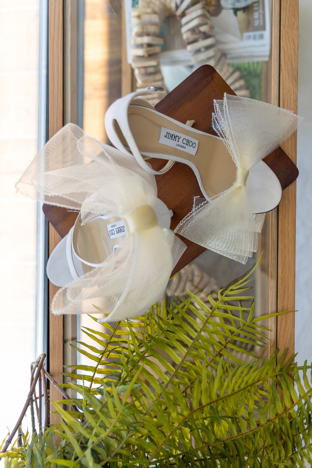 White bridal shoes with tulle bows displayed near a fern plant by a window.