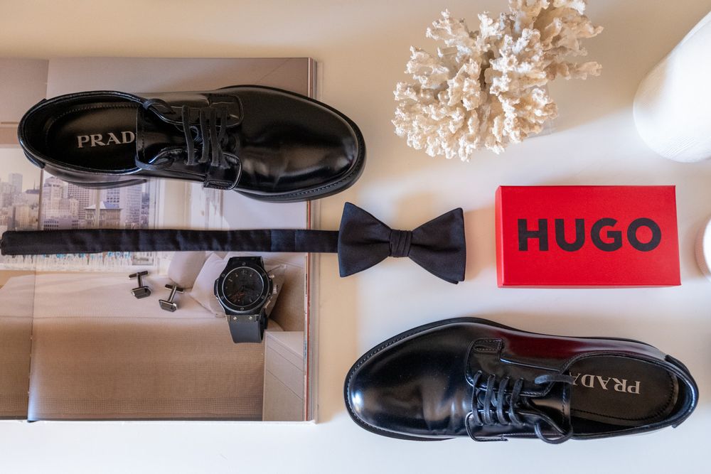 Wedding accessories including black dress shoes, bow tie, watch and Hugo brand label laid flat.