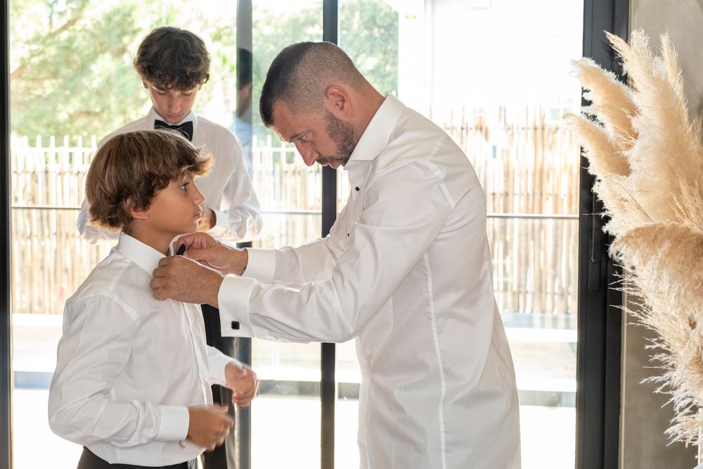 People in white shirts preparing formal attire by window with pampas grass decor.