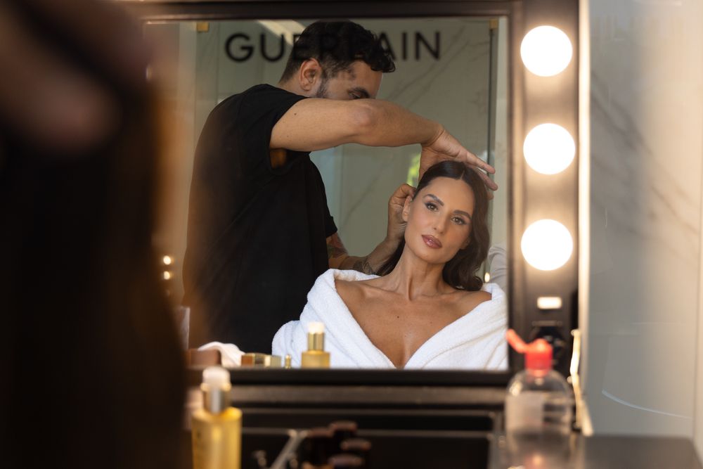 Reflection in vanity mirror shows styling session with Guerlain products visible.