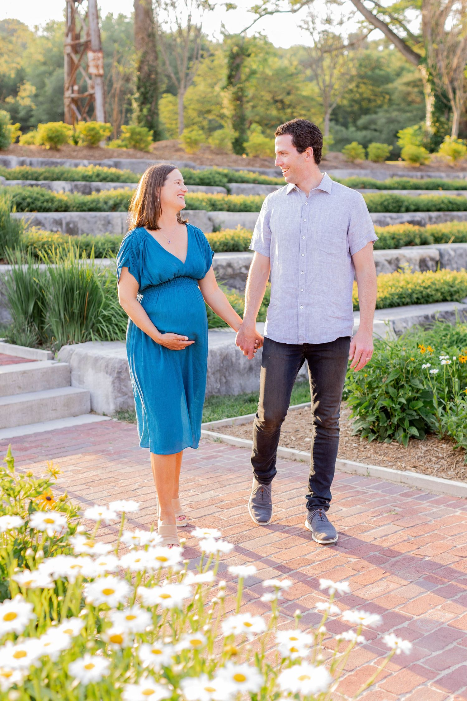 Couple embraces under wooden gazebo during golden sunset in nature setting with pregnant woman wearing blue dress.