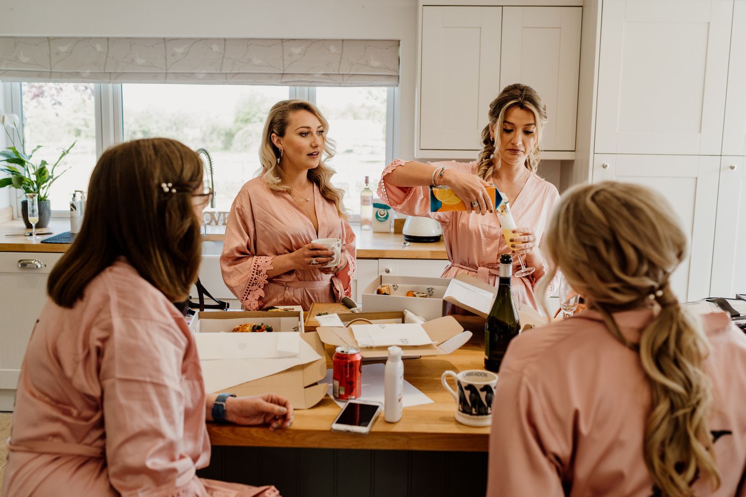 Multiple bridesmaids in pink robes gathered in a bright kitchen during wedding preparations.