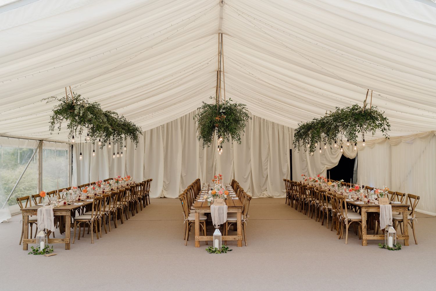 Elegant white wedding marquee interior decorated with hanging greenery and wooden chairs.