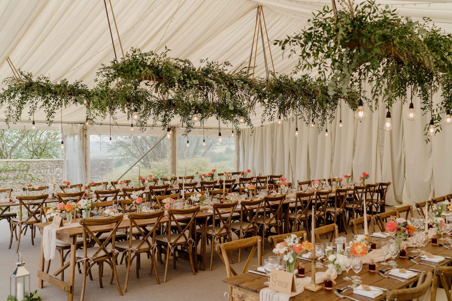 Long rustic wooden dining tables set beneath a white tent with hanging greenery and string lights create an elegant reception space.