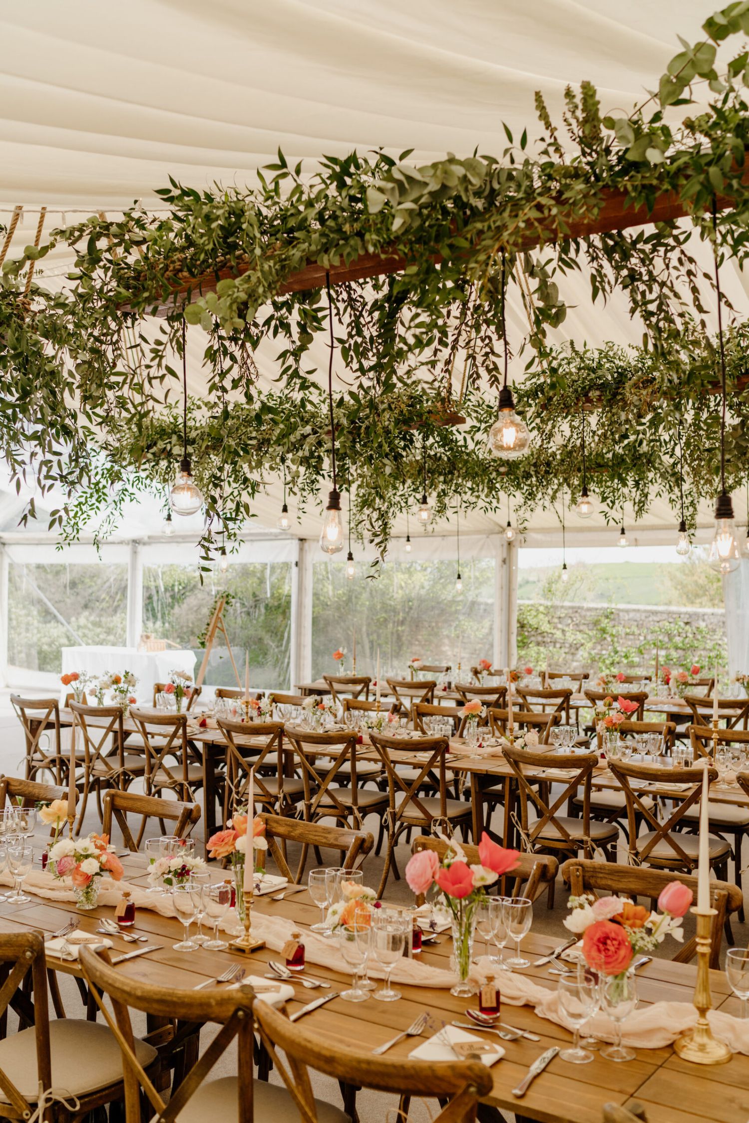 Reception tables arranged with pink floral centerpieces and place settings under a tent draped with natural greenery.