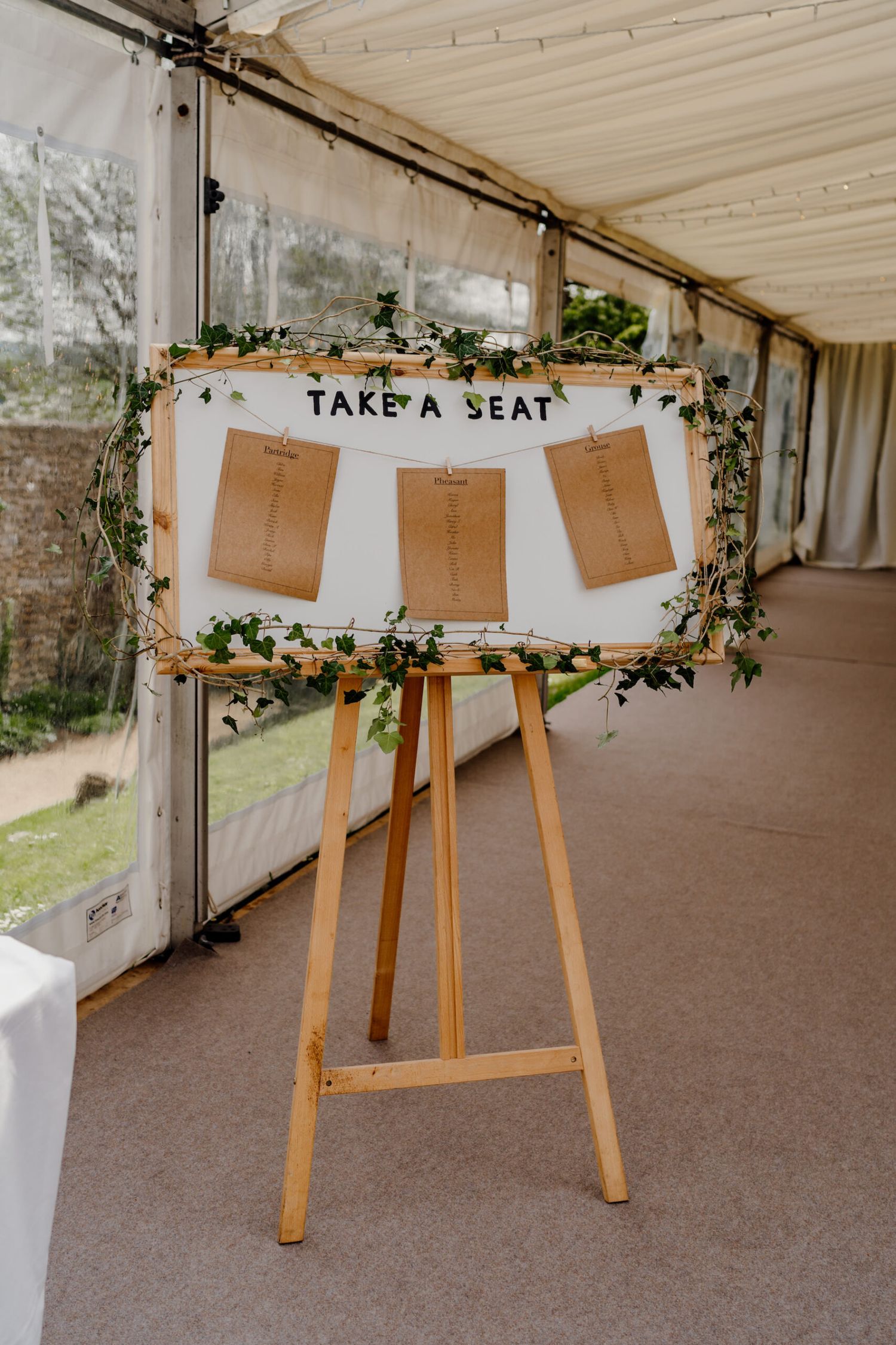 Wooden seating chart display with kraft paper cards hanging from string under 'Take a Seat' sign.