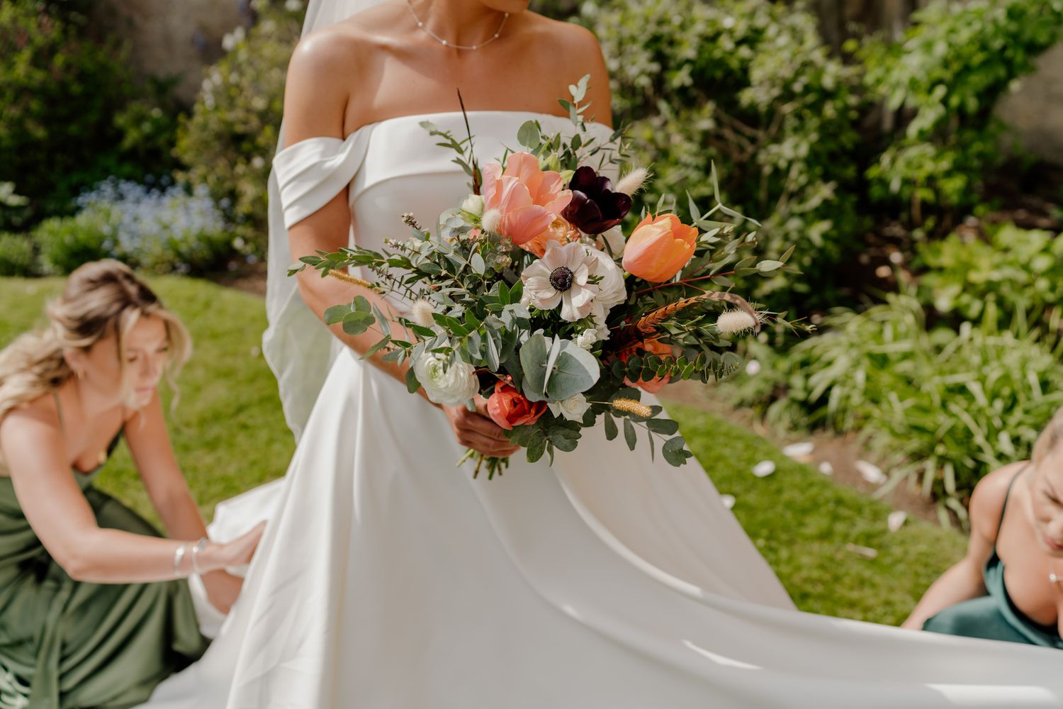 A wedding bouquet with coral and peach flowers being held against a white gown in an outdoor garden setting.