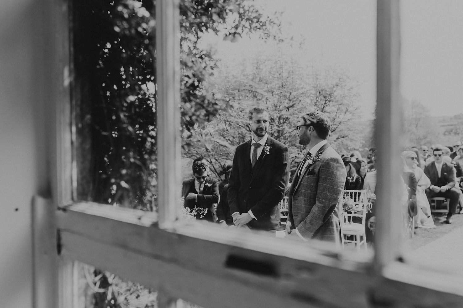 Black and white photo of wedding ceremony guests viewed through window panes.