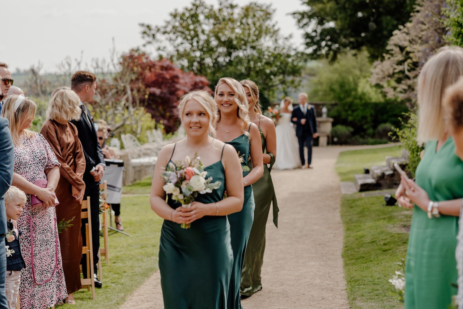 Wedding attendants in green dresses walking along garden path during ceremony.