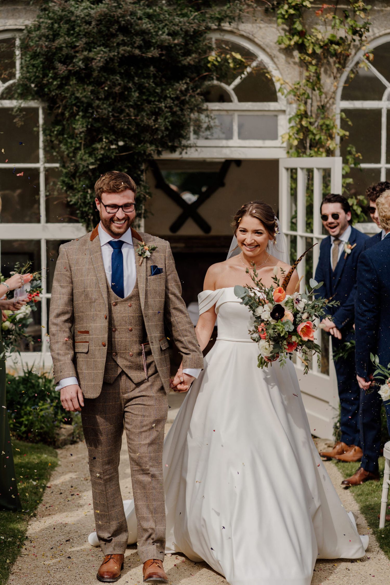 Wedding procession walking along garden path with stone building backdrop.