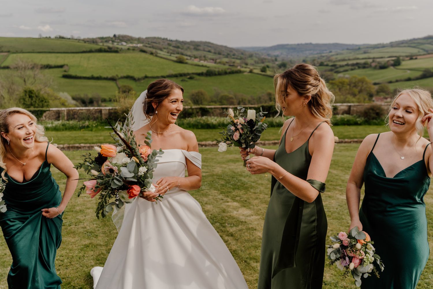 Wedding party in emerald green dresses posing against rolling countryside backdrop.