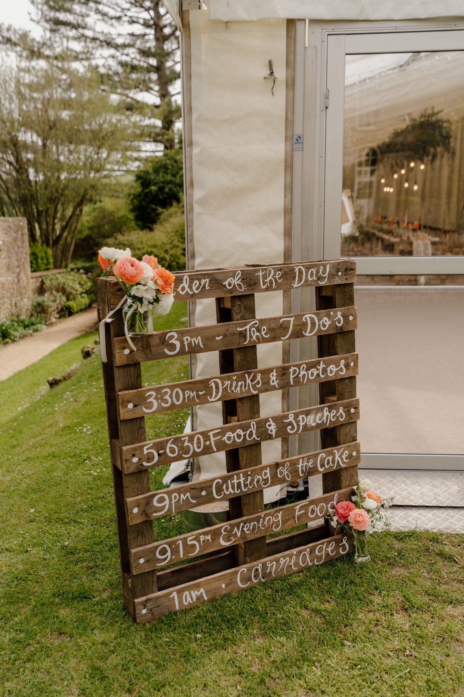 Wooden wedding timeline sign with floral decorations displays the day's schedule of events.
