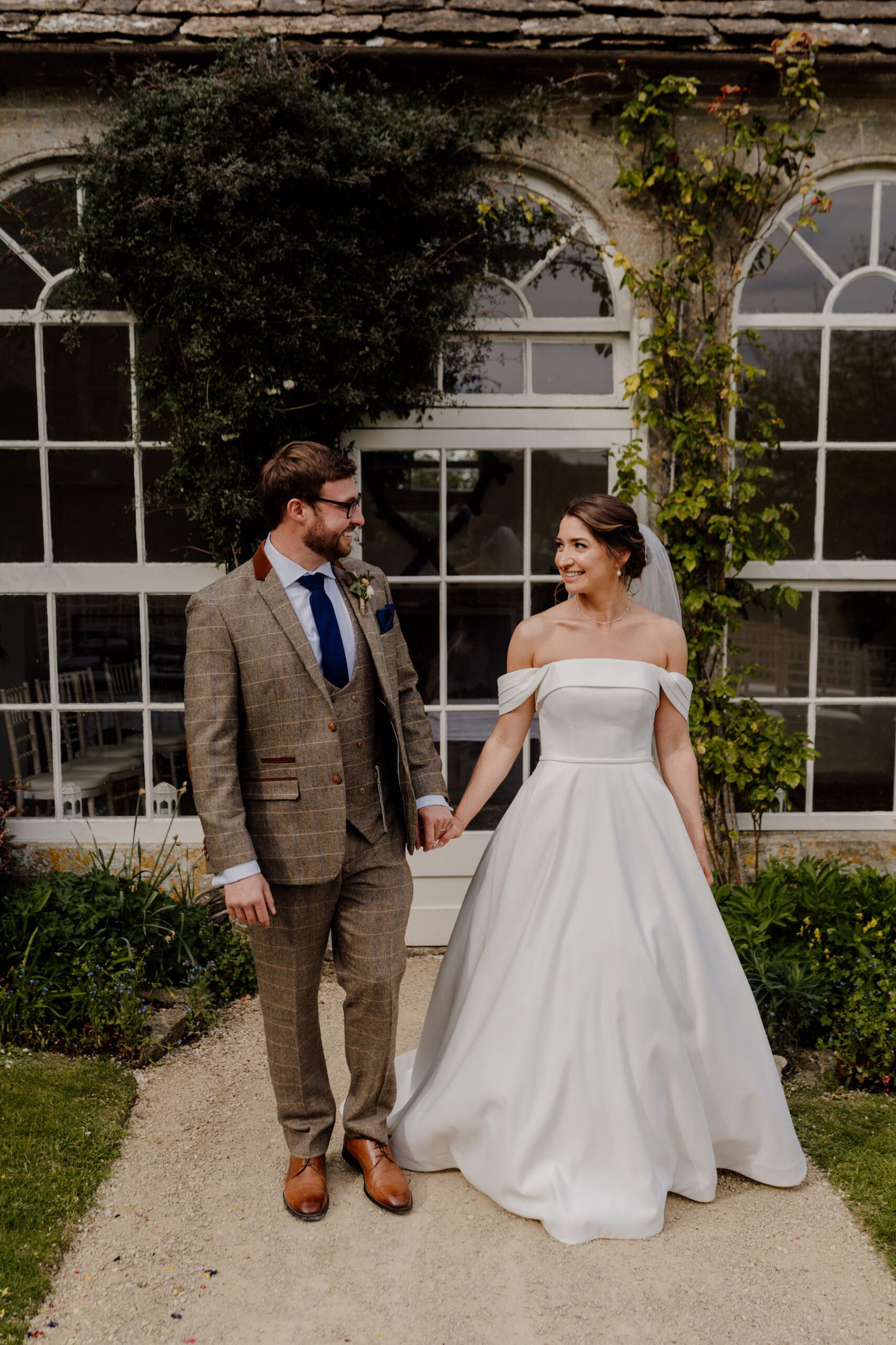 Couple in formal attire poses against historic building with ivy-covered windows.