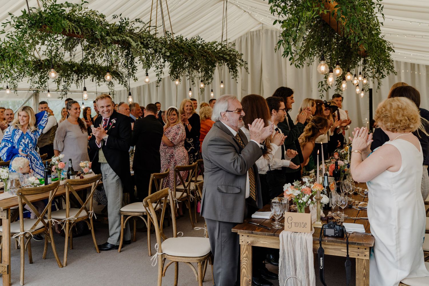 Wedding reception guests standing and applauding in an elegant tented venue with greenery decor.