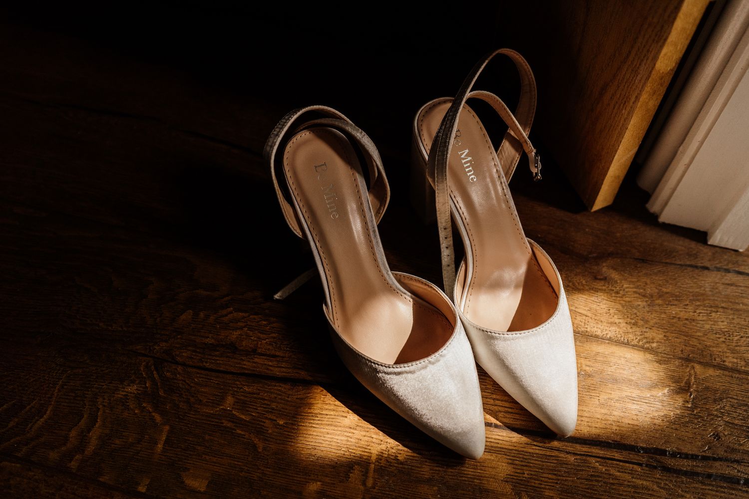 White bridal shoes sitting on dark wooden floor with dramatic side lighting.