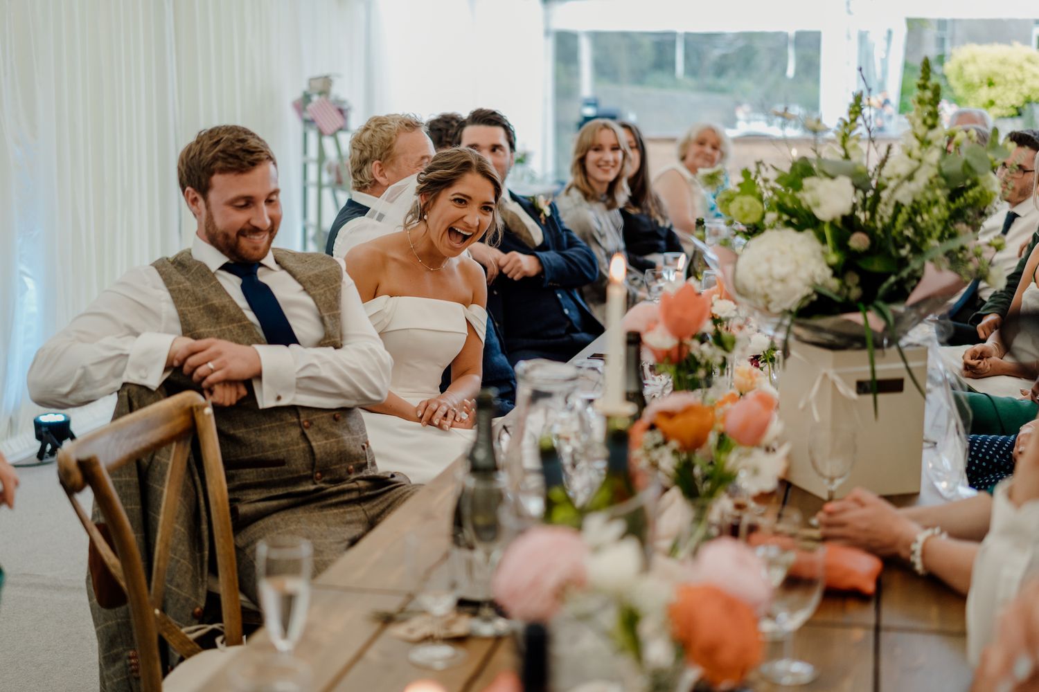 Wedding celebration with guests laughing at decorated reception table under marquee tent.
