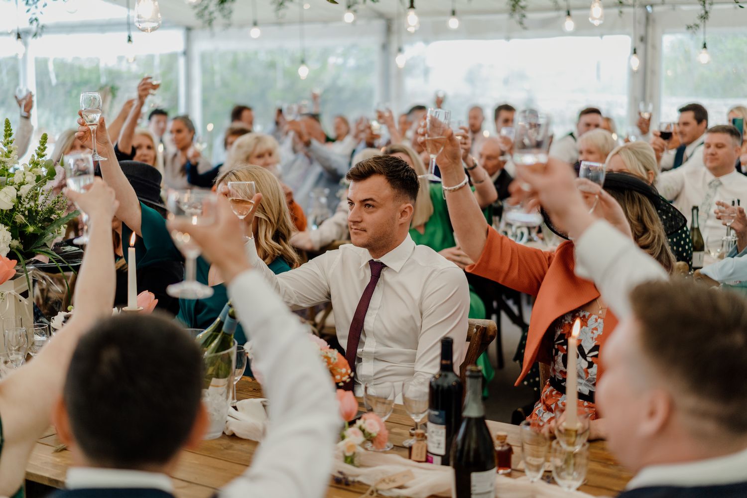 Wedding reception guests raise their hands in celebration under a white tent.