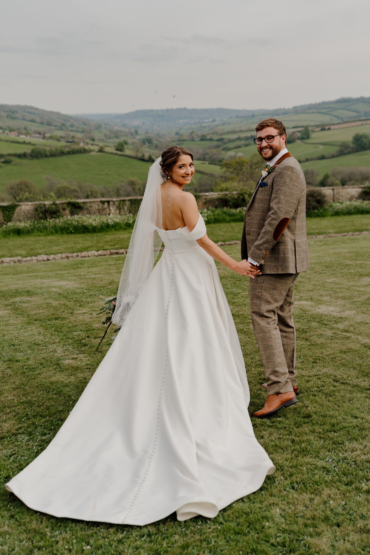 Couple poses on a grassy hilltop with a flowing white wedding dress against a scenic countryside backdrop.