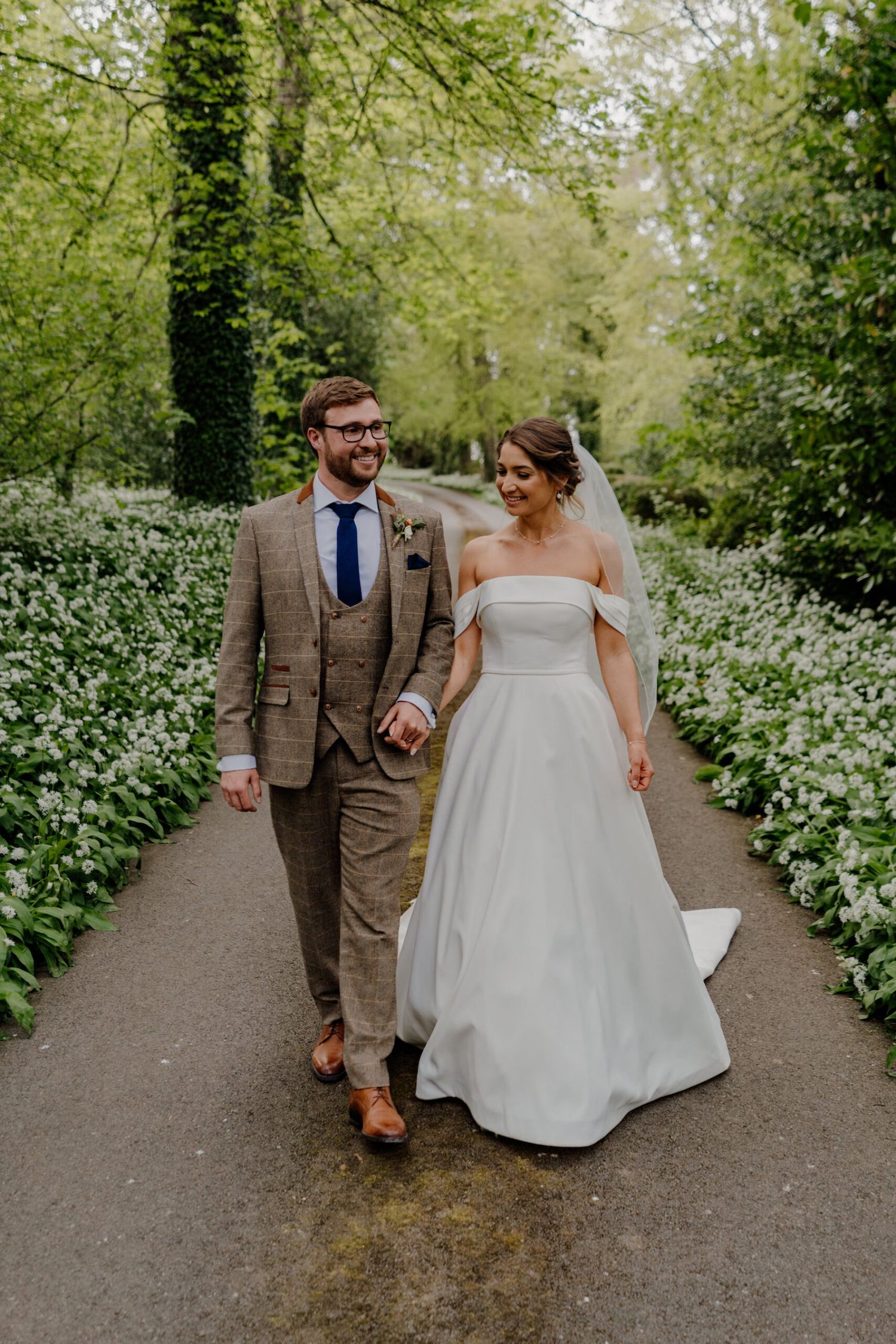 A newlywed couple walks along a garden path lined with white flowers and lush greenery.