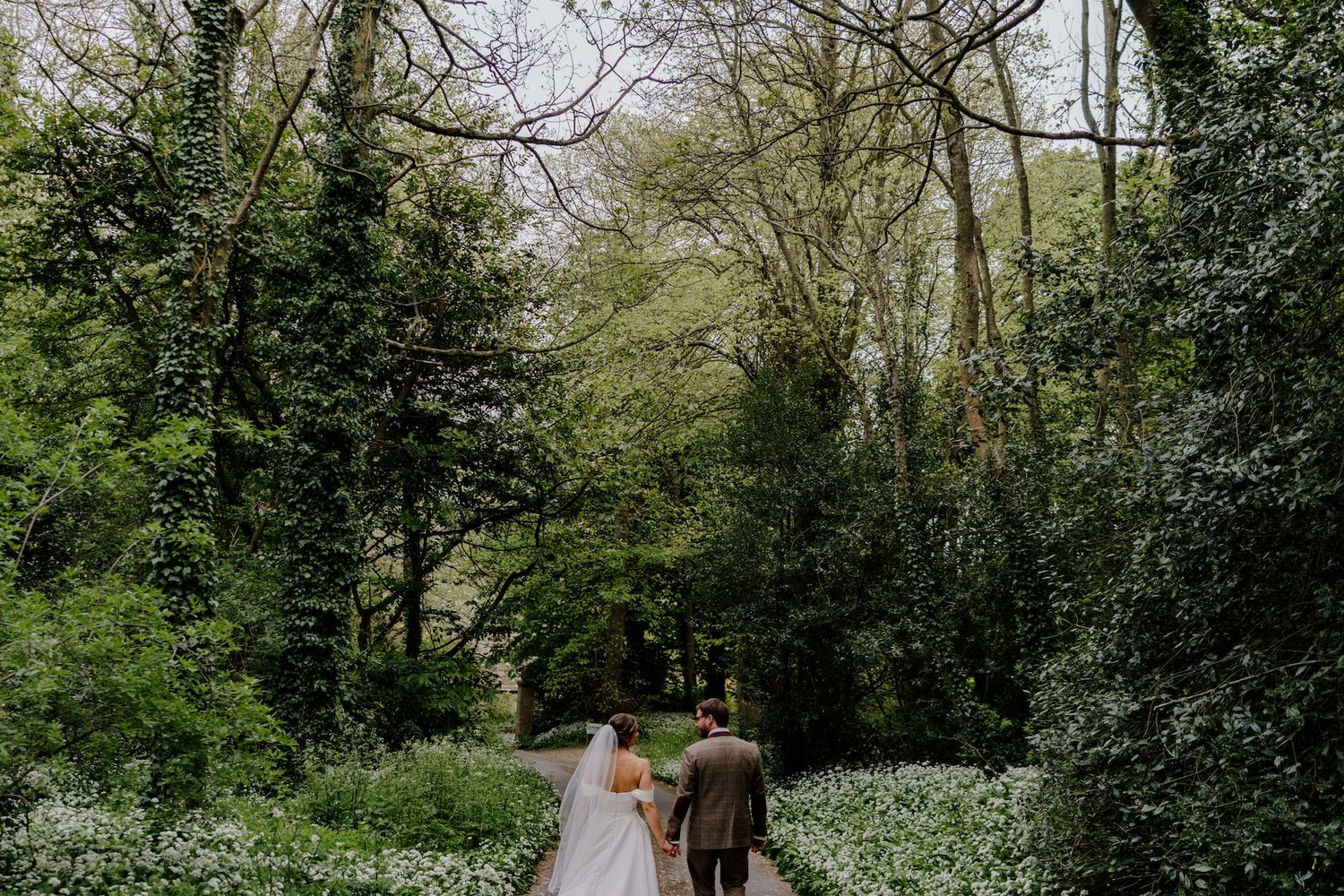 A couple in wedding attire strolls down a tree-lined path surrounded by white wildflowers.