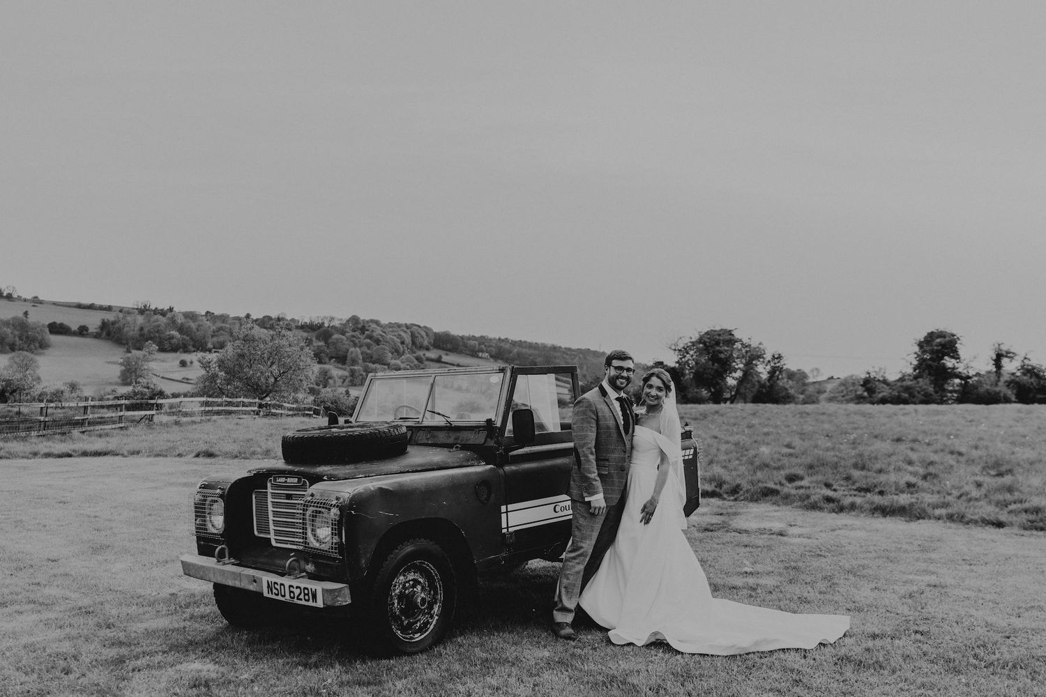 A classic Land Rover serves as a backdrop for a wedding photo in a rural setting.