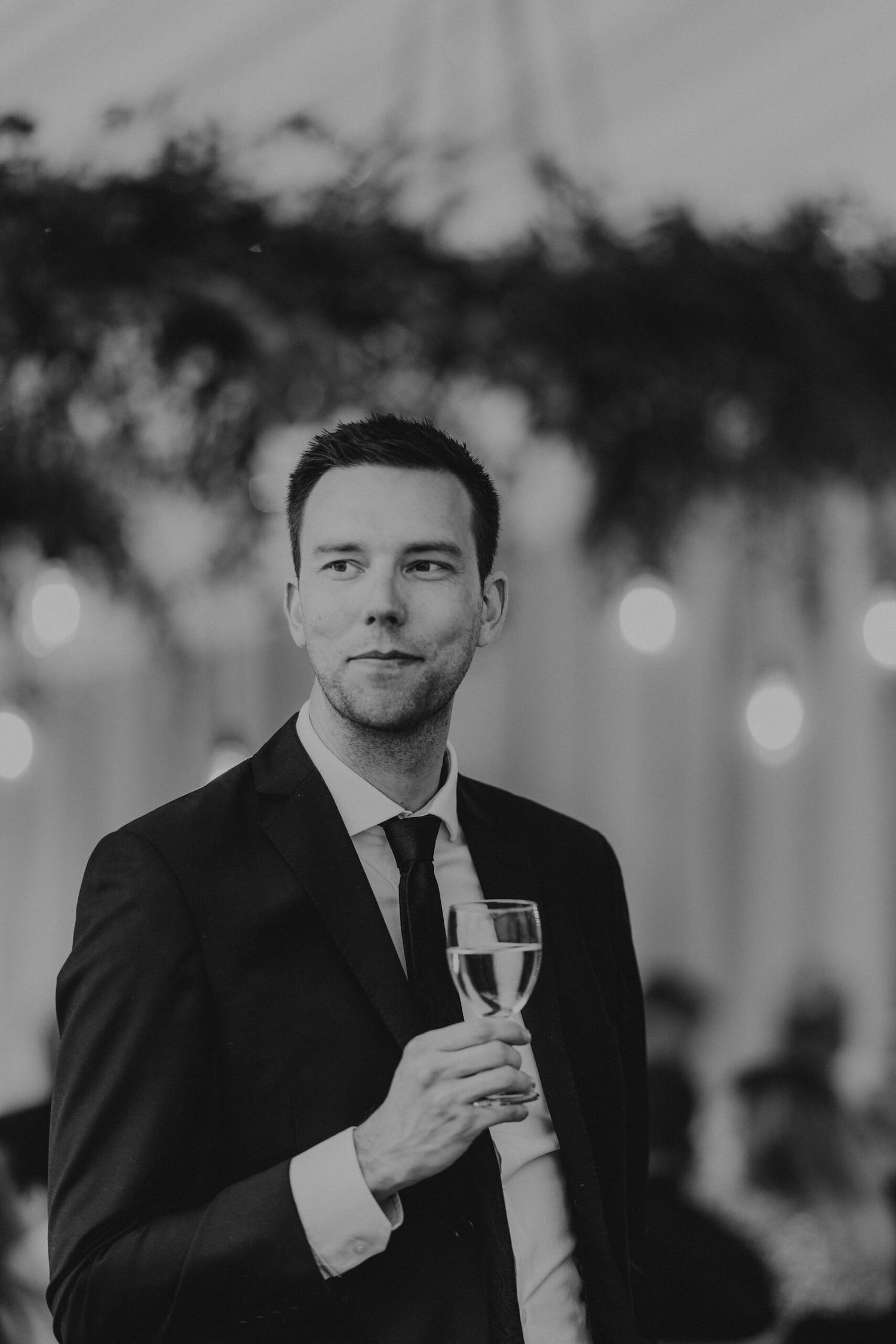 A formal wedding guest holds a champagne glass at an evening reception with bokeh lights.