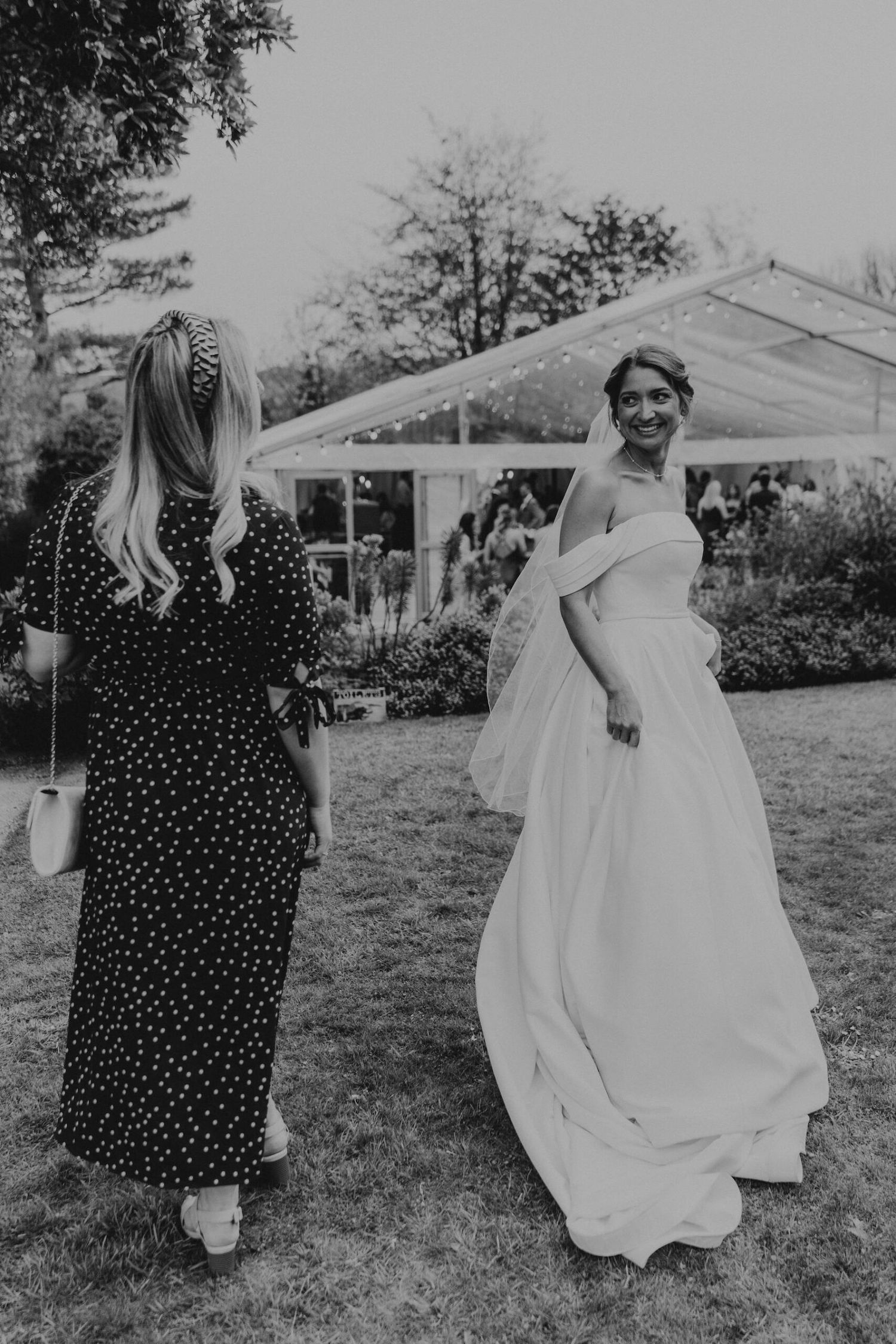 A bride in a flowing white gown twirls in a garden setting next to a guest in polka dots.