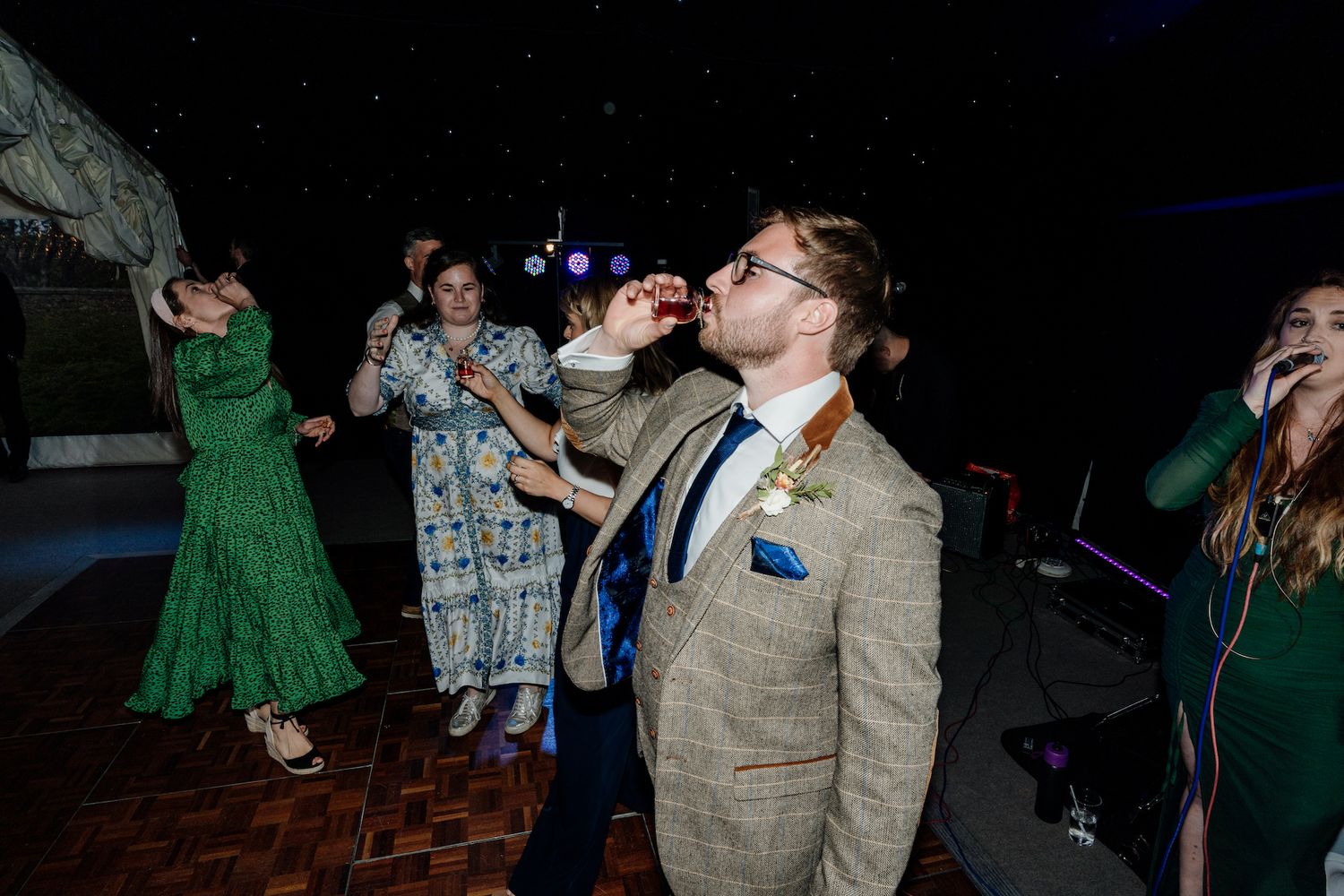 Wedding guests take shots on a dark dance floor during reception celebrations.