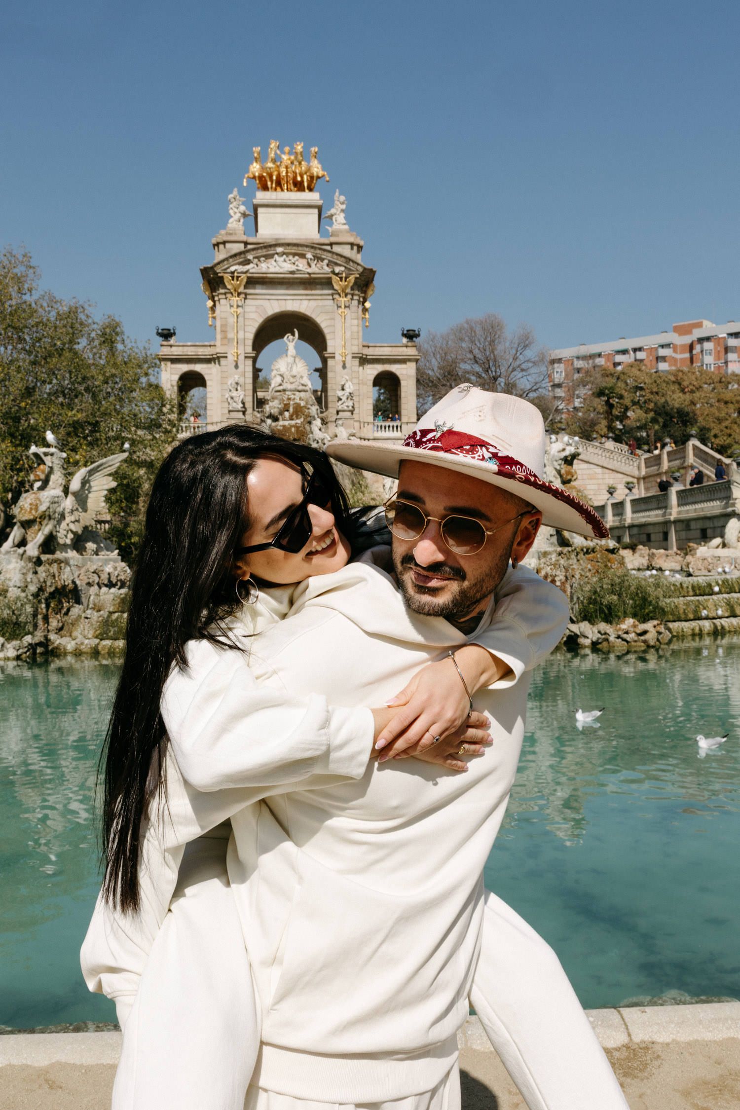 Couple in white outfits embracing near a decorative fountain with classical architecture.