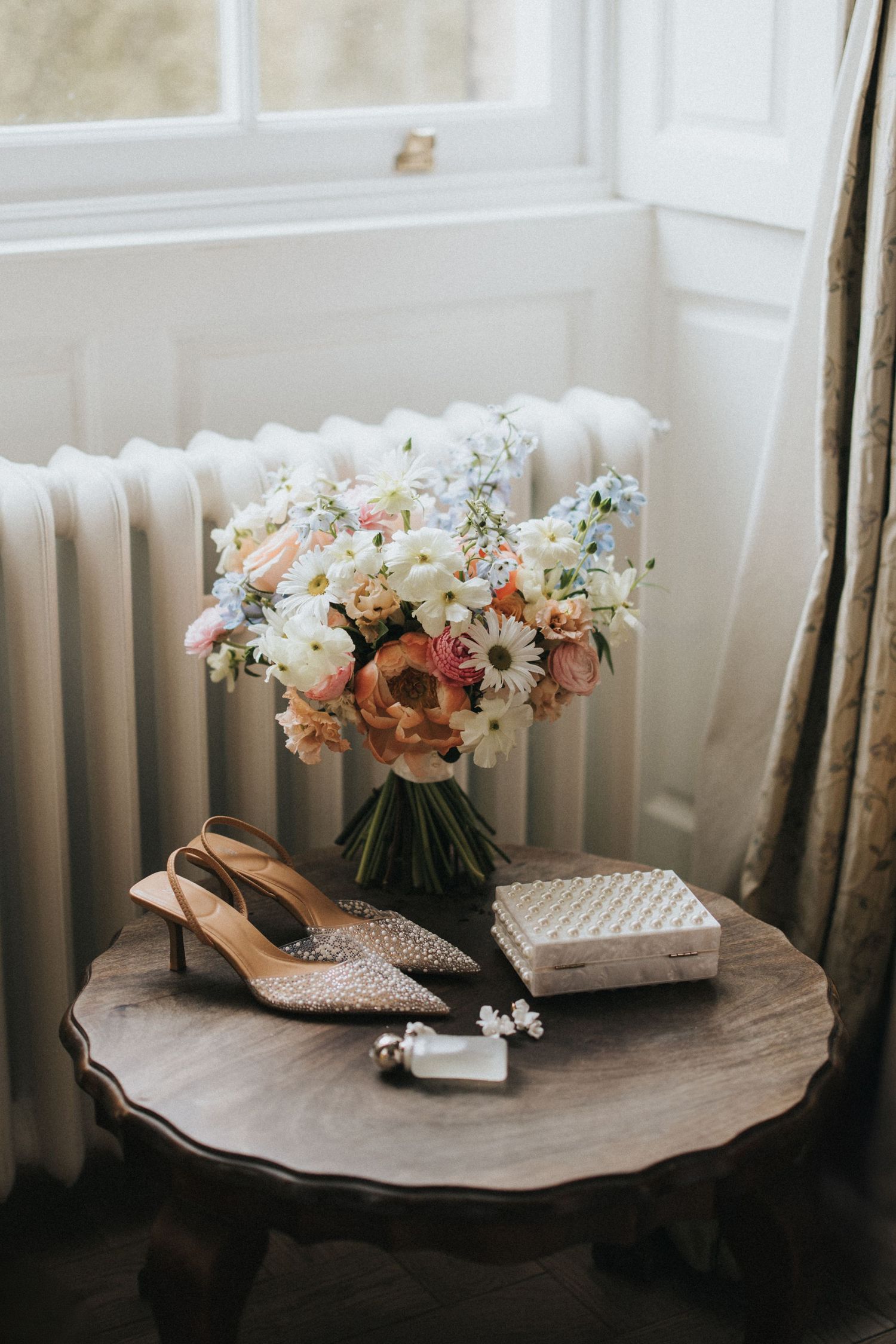 Wedding details with sparkly bridal shoes, floral bouquet in pastel coloyrs, and jewellery on vintage wooden table by window.