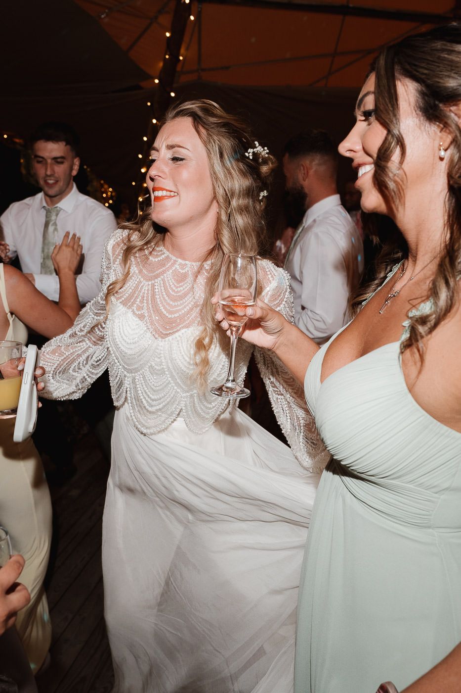 Wedding guests celebrate and dance at a reception with string lights overhead, wearing white and mint green dresses.