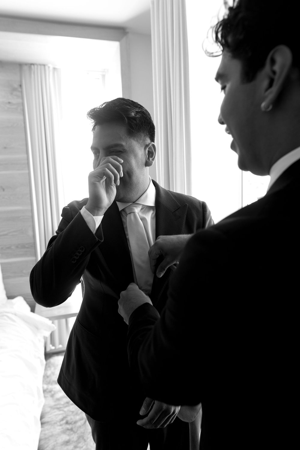 Two men in formal black suits share an emotional candid moment near a window in a bright indoor setting.