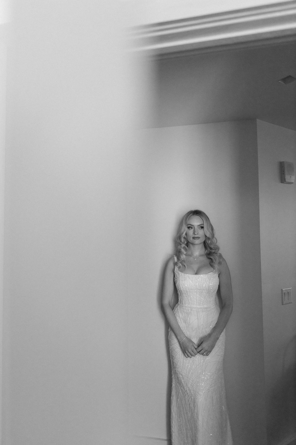 A black and white portrait of a bride in a fitted wedding dress standing in a minimalist interior.