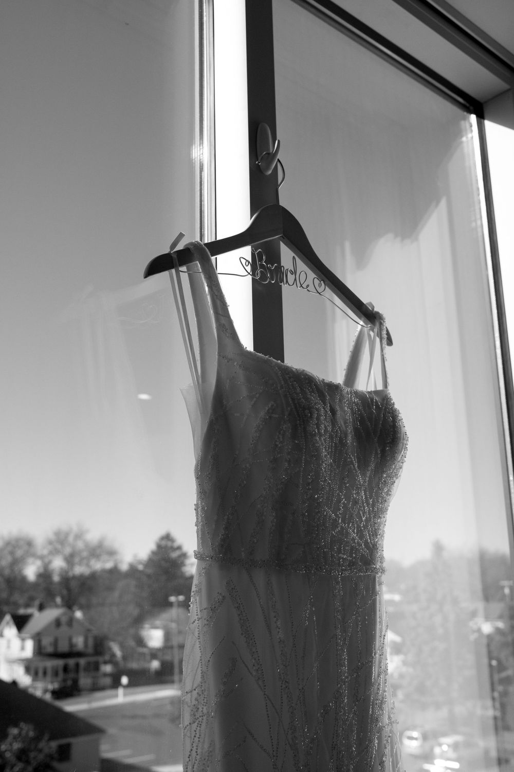 A black and white image of a delicate wedding dress hanging on a St. Puccis hanger in front of a window.